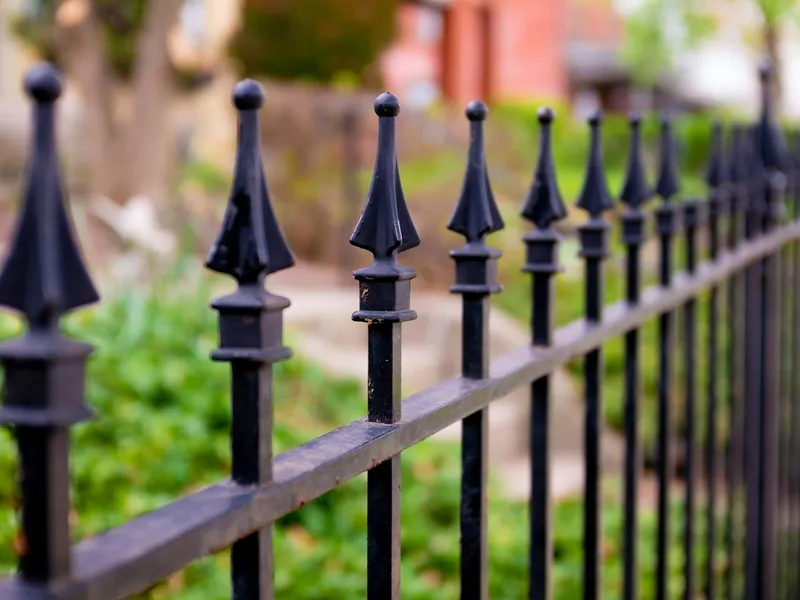 Close view of the cast spear finials along the top of a black steel railing fence, the line of them receding into soft focus