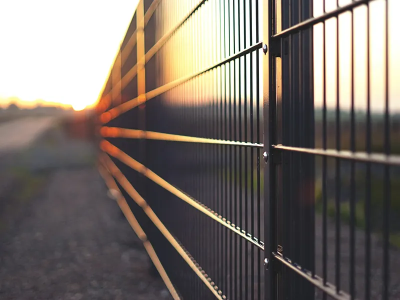 A line of welded mesh security fencing on steel posts running away towards a low sun, seen close along the face of the panels